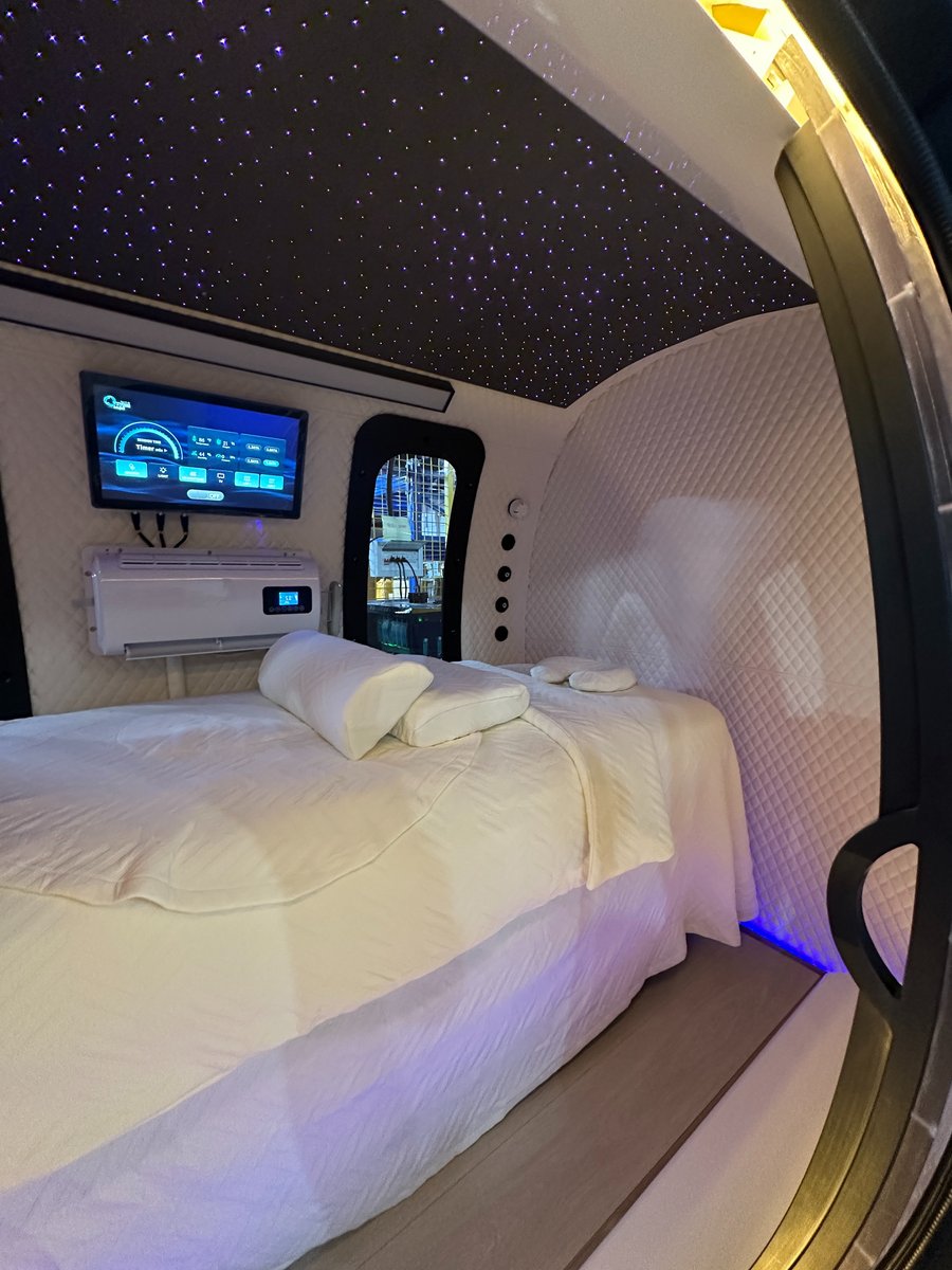 Angled view of the ExS64 sleep configuration showing the quilted curved walls, bed, touchscreen, air-conditioning unit, arched window, and starlight ceiling