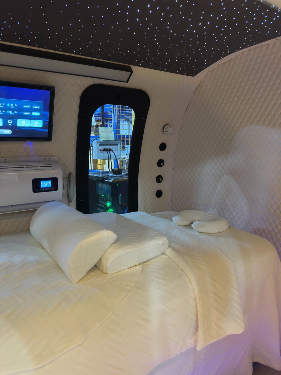 ExS64 interior in the sleep configuration — a wide white quilted bed with pillows beneath a fiber-optic starlight ceiling, a wall-mounted touchscreen, air-conditioning unit, and arched viewing window