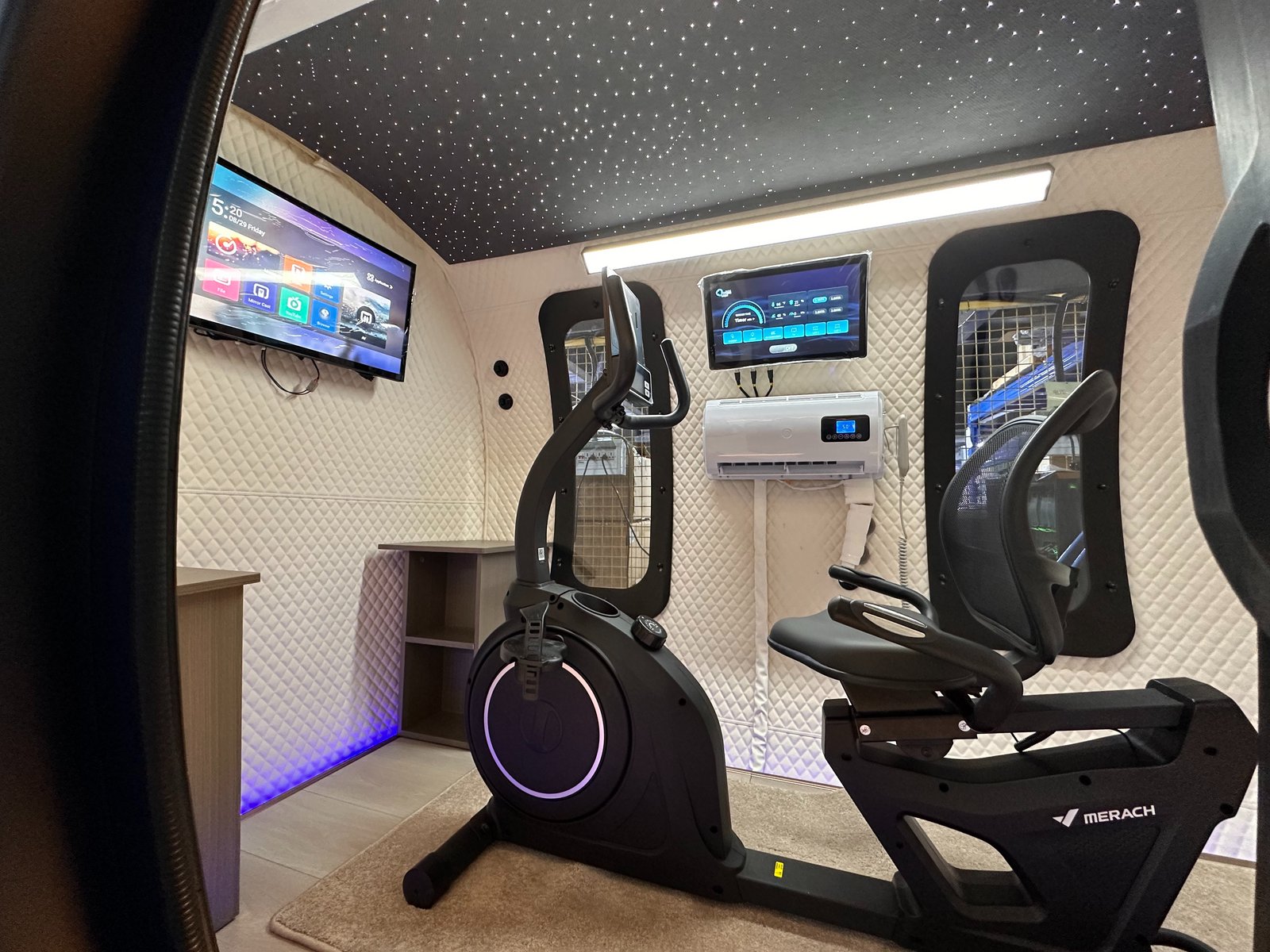 ExS64 exercise configuration with a MERACH recumbent bike and upright bike, wall-mounted TV, touchscreen control, air-conditioning unit, and starlight ceiling