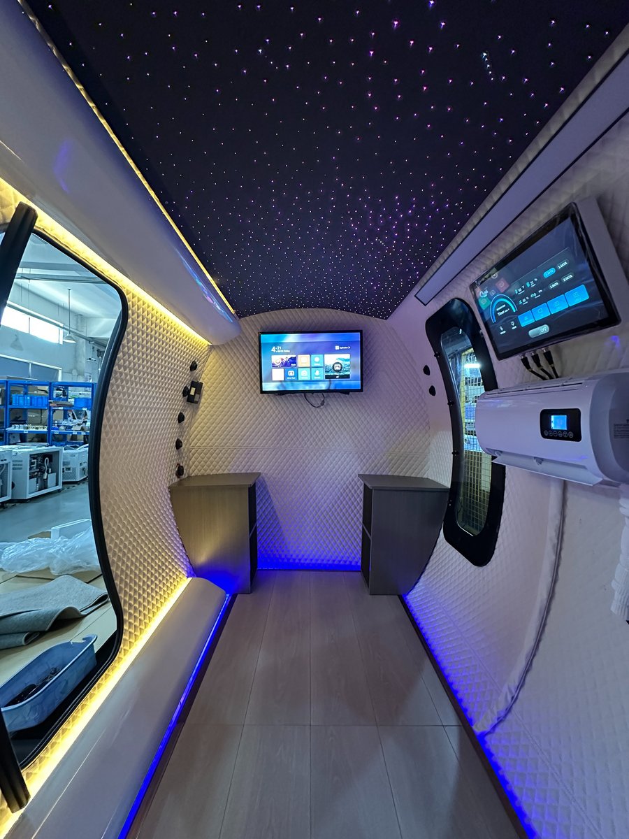 Walk-in corridor view of the ExS64 with quilted white walls, starlight ceiling, wall-mounted TV, touchscreen control, air-conditioning unit, dark wood side benches, and blue and warm LED accent lighting