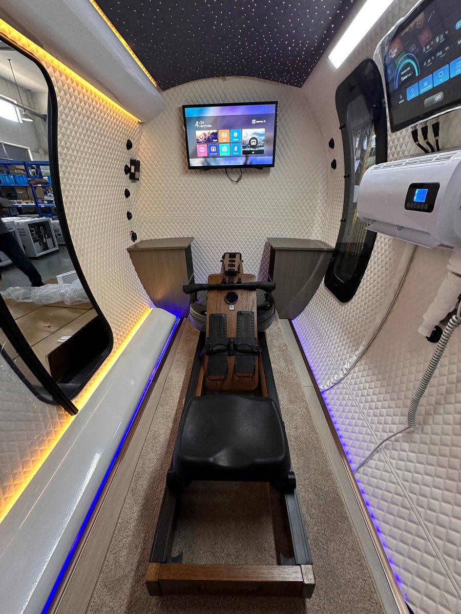 ExS64 exercise configuration with a wooden rowing machine centered on the floor, wall-mounted TV, touchscreen control, air-conditioning unit, mirror, and side benches